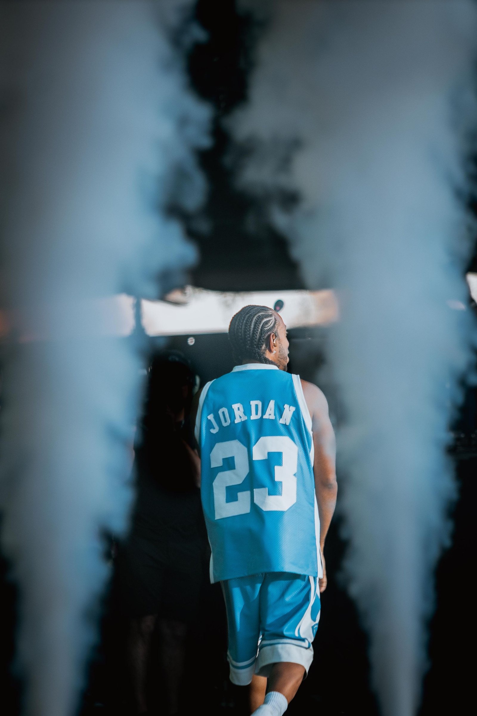 A performer in a light blue number 23 jersey stands between columns of stage smoke. Photograph by Gabe Key.
