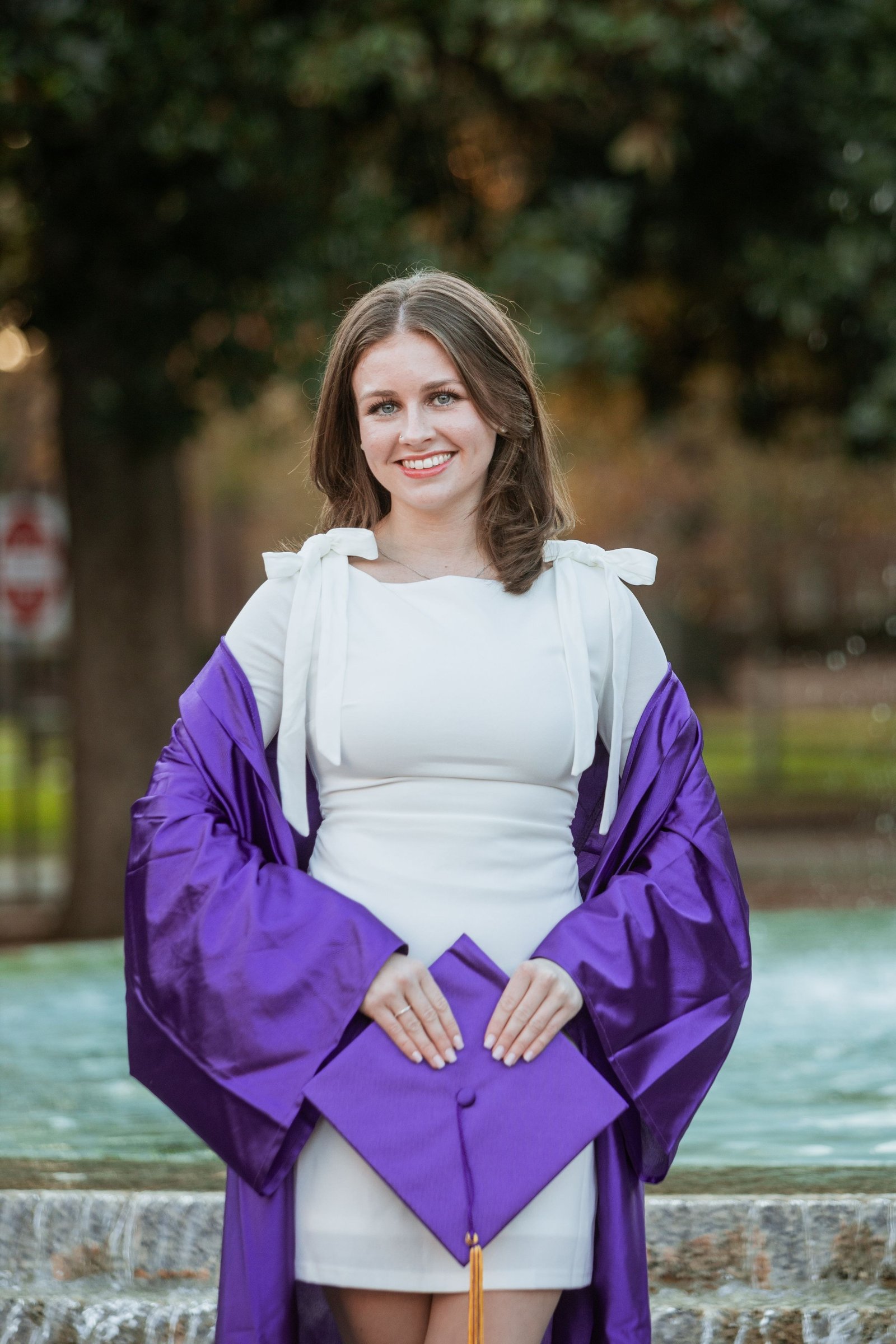 A graduate in a purple gown beside a fountain.