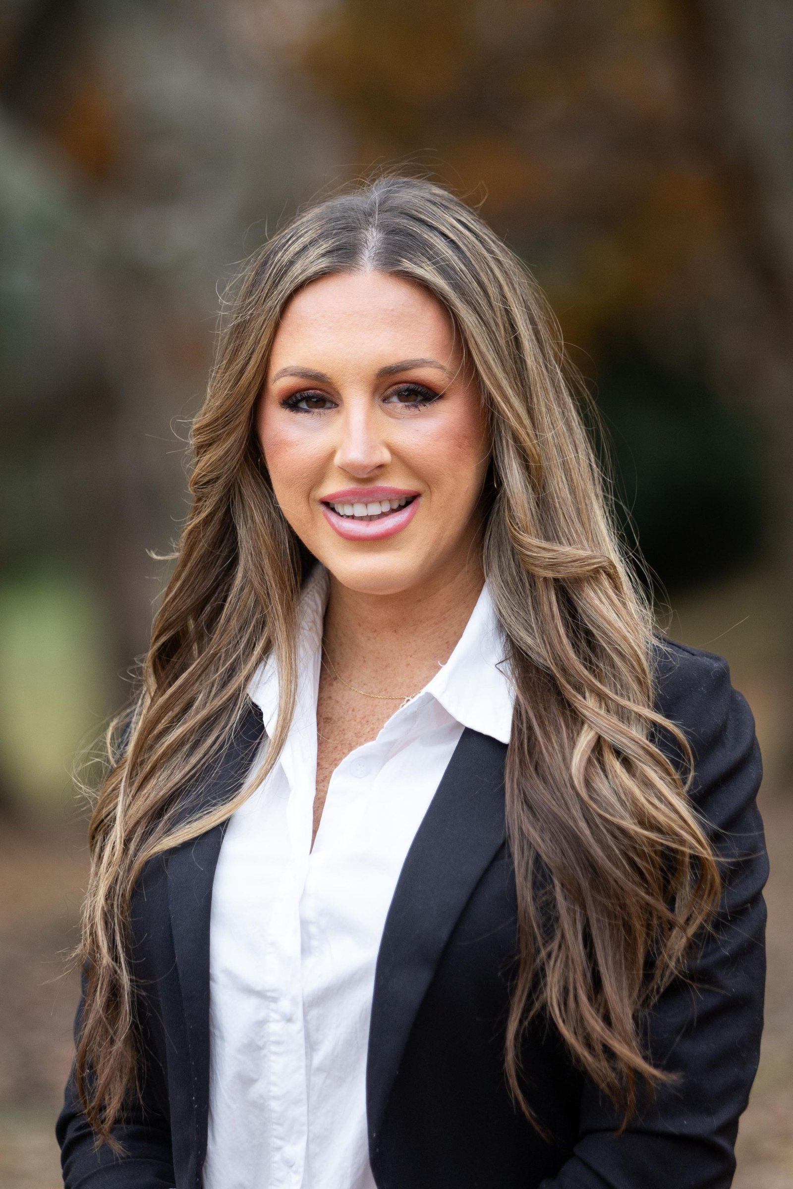 Professional headshot of a woman in a dark blazer, outdoors in autumn.